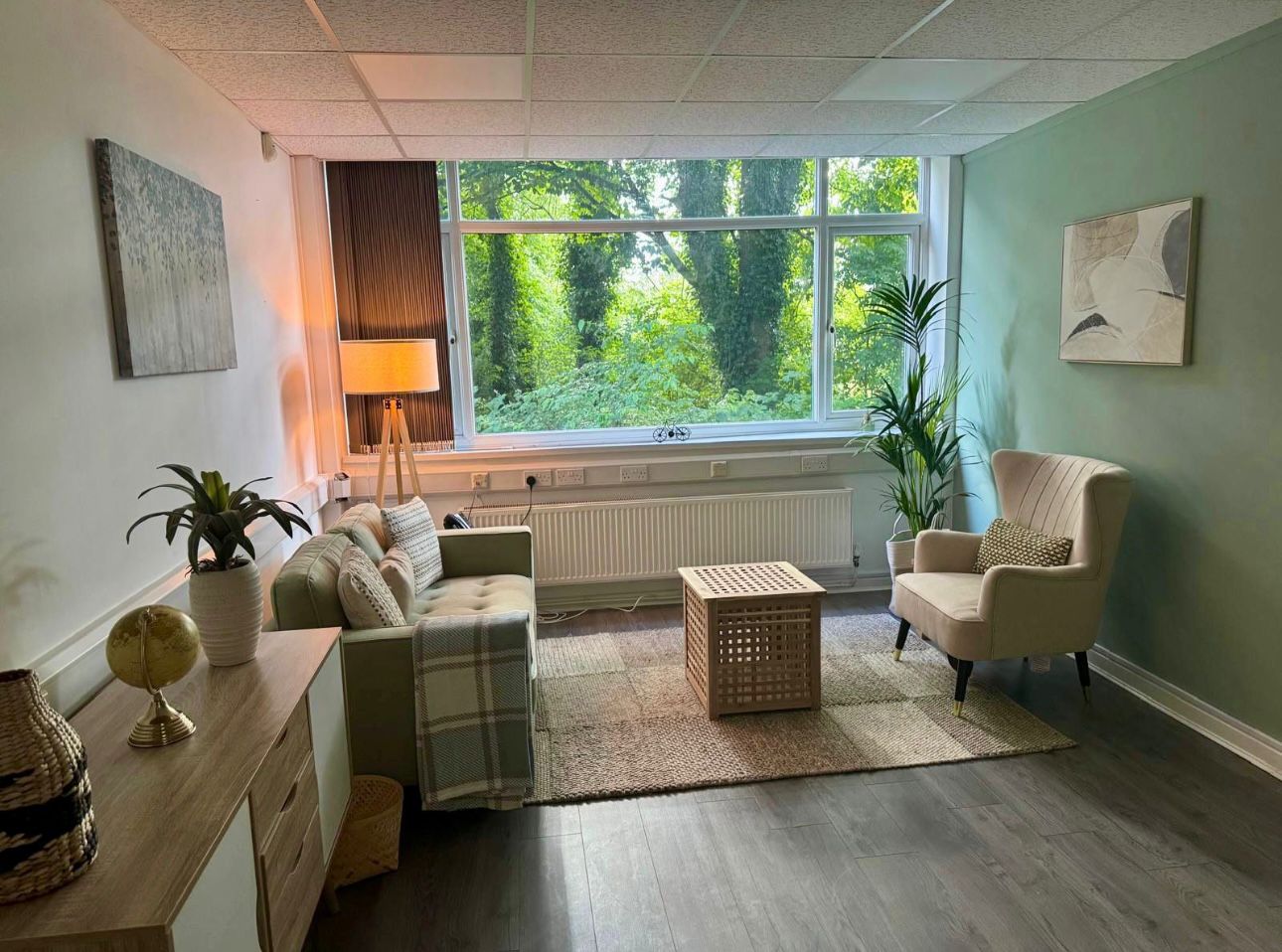 TC Psychology clinic room with a sofa, armchair, plants and a large window overlooking trees